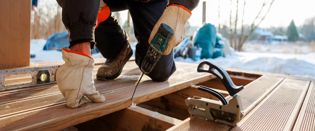 Protecting Your Deck From Hail in Eastern Nebraska A contractor in work gloves using a power drill to perform a deck repair on a wooden patio during winter.