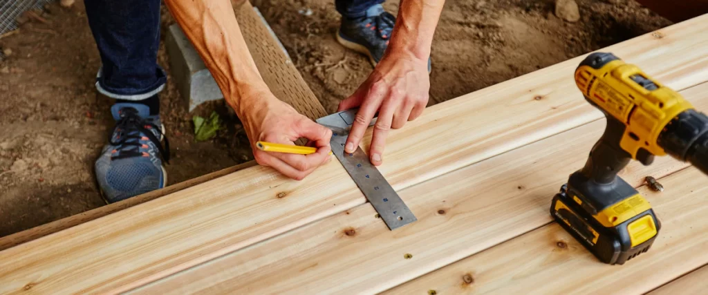 Deck Design for Pole Barn Homes & Shouses in Nebraska Close-up of a carpenter using a speed square and pencil to mark measurements on new wooden deck boards during construction.