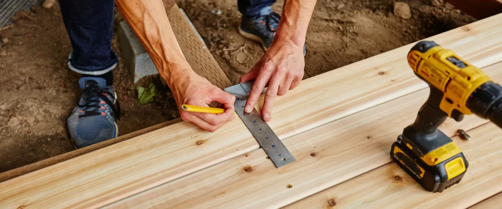 A close-up shot of a contractor's hands using a speed square and pencil to mark a cedar wood board for a custom deck installation project.