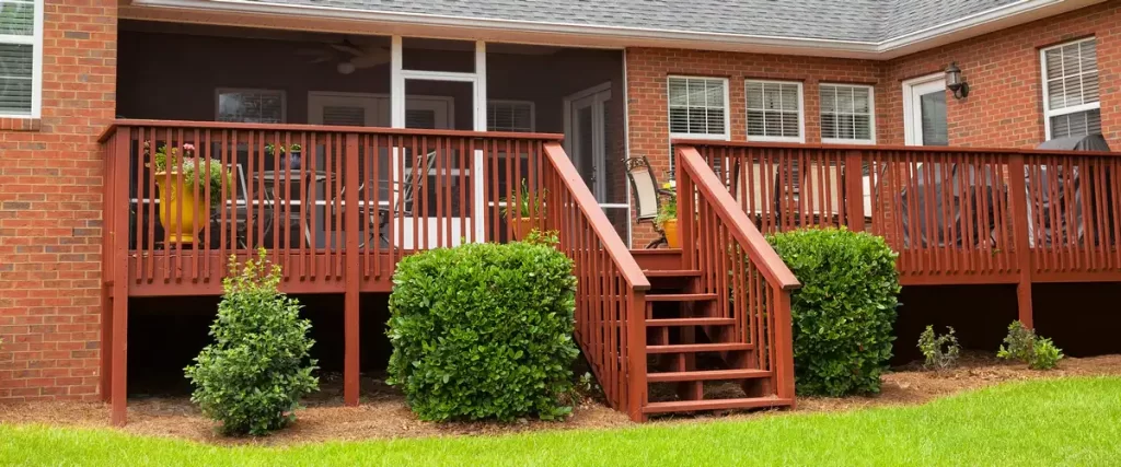 Deck Design for Pole Barn Homes & Shouses in Nebraska A reddish-brown stained wooden deck attached to a red brick house featuring stairs leading down to a lawn with manicured green bushes.