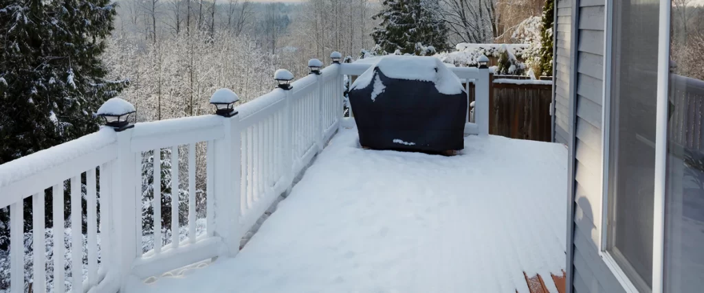 A white-railed residential deck completely covered in a thick layer of winter snow, showing a covered grill and solar post cap lights.