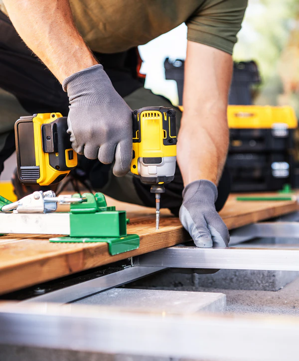 A worker wearing grey gloves uses a yellow power drill to securely fasten a wooden board, highlighting professional deck maintenance plans in Omaha, NE.