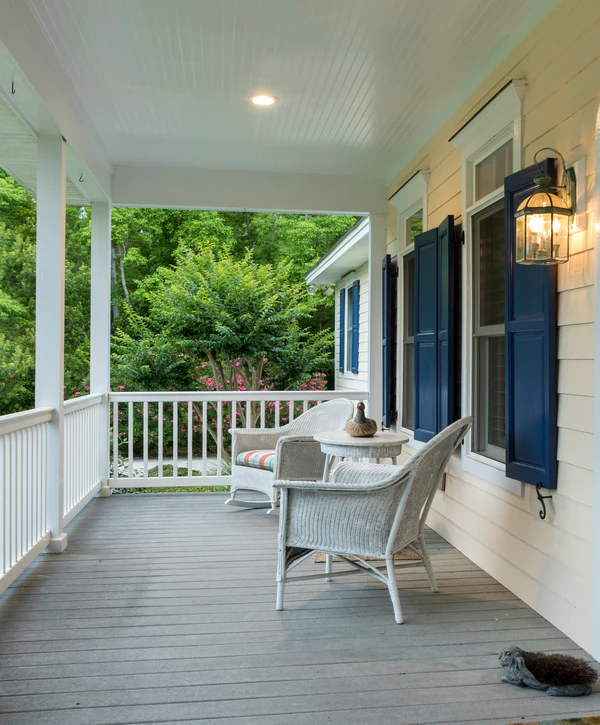 A charming covered porch and Deck Building project in Fort Calhoun, NE, featuring grey painted flooring, white wooden railings, white wicker chairs, and dark blue house shutters.