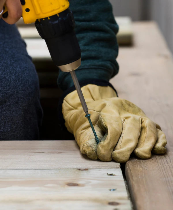 Close-up of a worker wearing heavy leather gloves using a power drill to drive a screw into wooden boards during a deck repair in Chalco, NE.