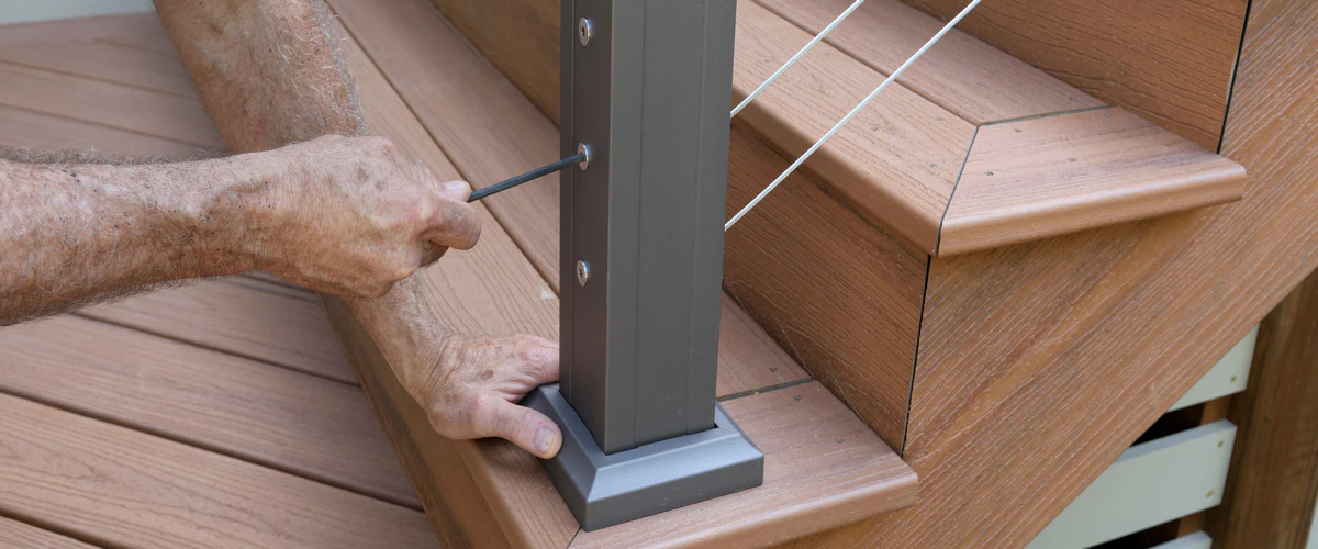 Close-up of a worker's hands using a hex key to tighten the hardware on a metal railing post for a composite deck repair.