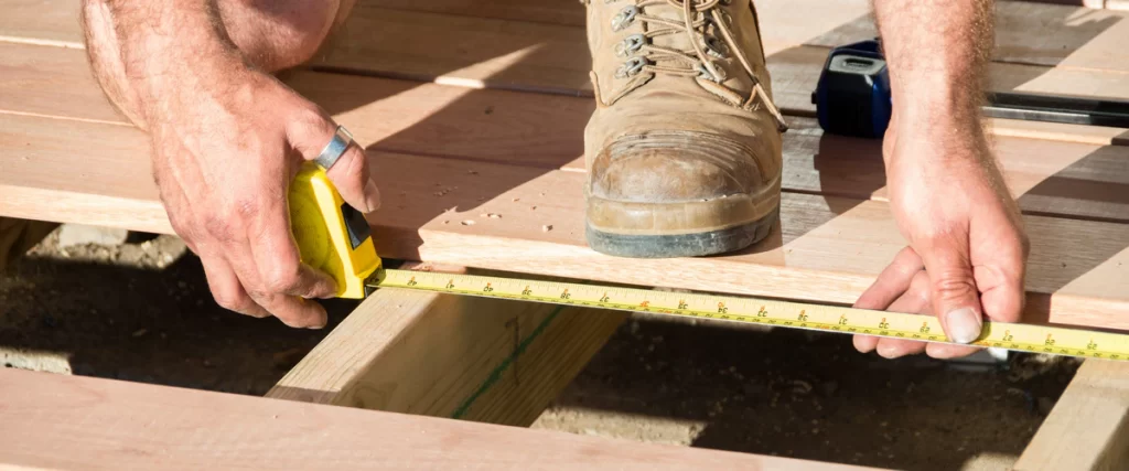 Worker wearing heavy boots performing a deck inspection by extending a tape measure across the wooden floor joists.