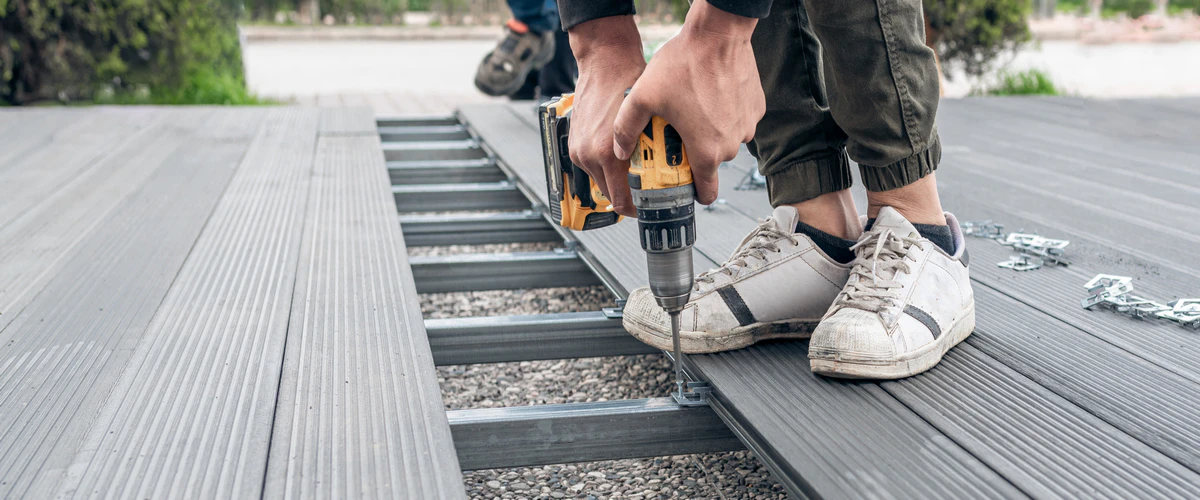 A professional executing a composite deck repair by using a power drill to install metal hidden fasteners between grey decking boards over a steel frame.
