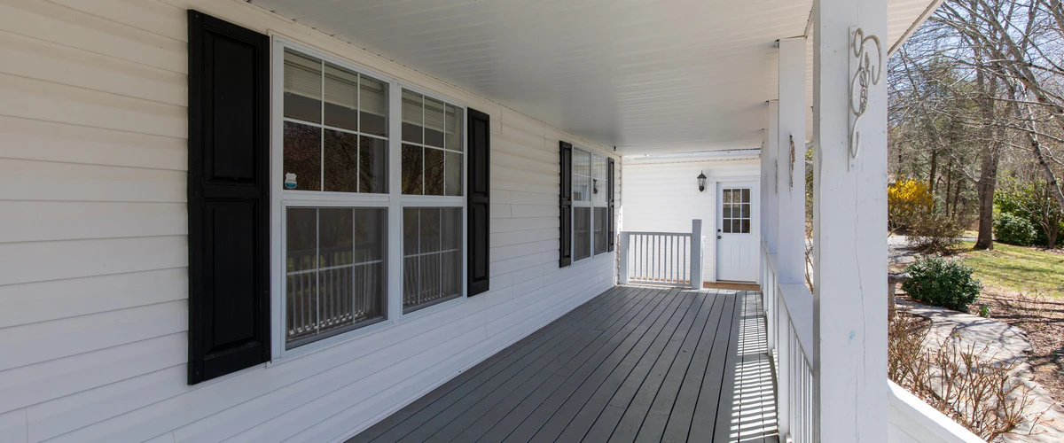 Covered Deck Builder in Council Bluffs, IA A long residential covered deck attached to a white house with black shutters, featuring a grey painted wooden floor and traditional white railings leading to a front door.