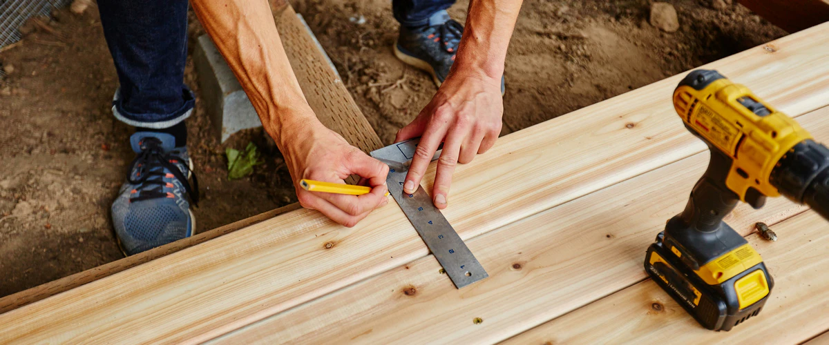 A contractor uses a metal square ruler and pencil to carefully measure and mark wooden boards alongside a power drill for a precise deck repair.