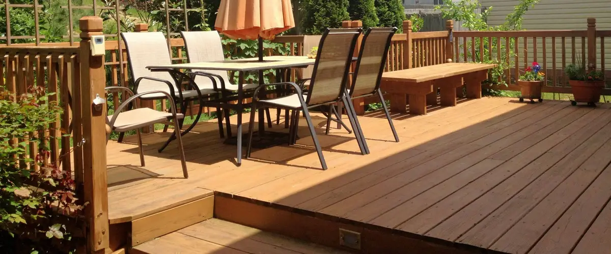 A multi-level backyard wood deck equipped with a dining table, a closed orange umbrella, chairs, traditional wooden railings, and a custom built-in wooden seating bench.