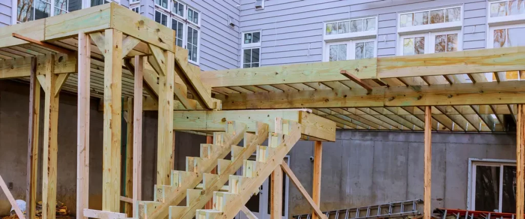 A robust wooden deck structure under construction attached to a house, featuring exposed joist framing, support posts, and stair stringers.