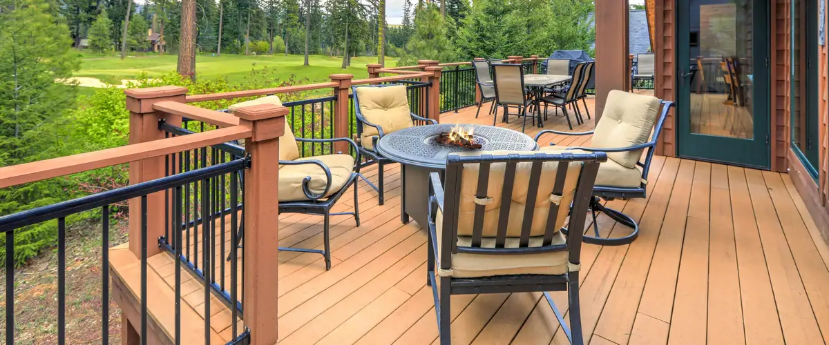 A spacious outdoor wooden deck featuring a round fire pit table, tan cushioned chairs, and black metal railings with a scenic background view of a golf course and pine trees.