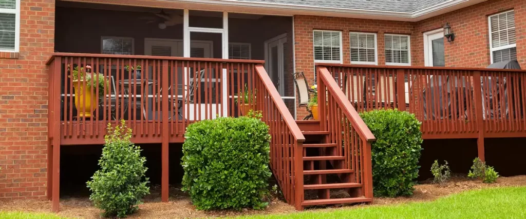 Red-stained wooden deck stairs leading up to a large backyard deck attached to a brick house, surrounded by green landscaping bushes.