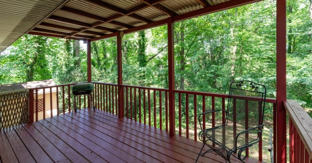 A red painted wood patio constructed by a Covered Deck Builder in Council Bluffs, IA, featuring a translucent corrugated roof, a green barbecue grill, and a black iron chair facing dense green woods.