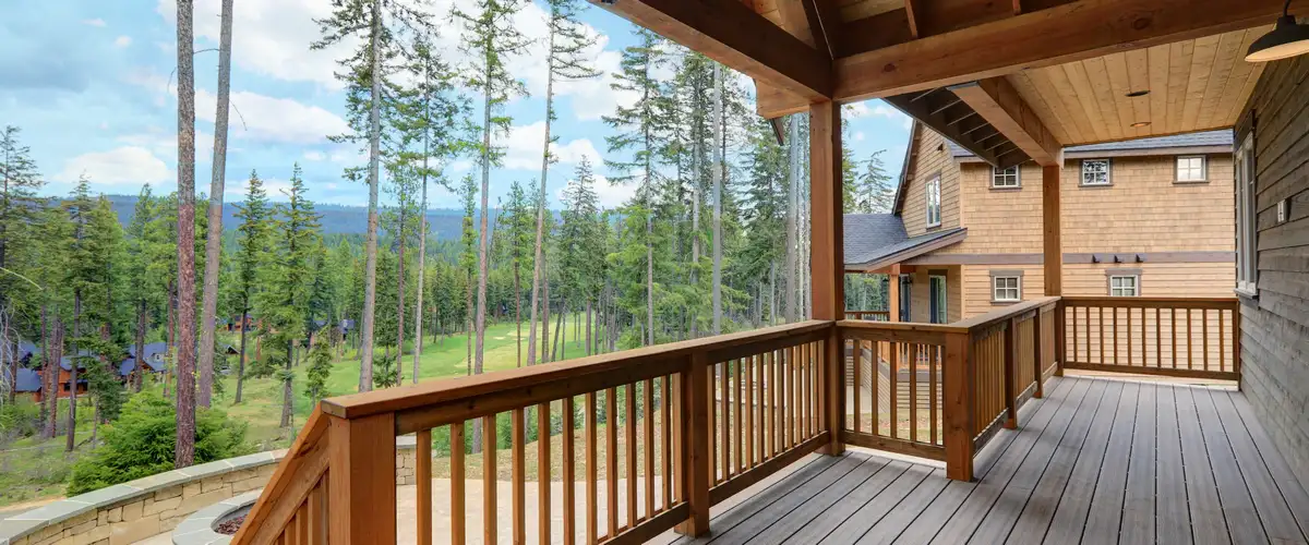 Covered Deck Builder in Council Bluffs, IA A rustic covered deck on a wood-sided home featuring grey flooring and thick wooden railings, looking out over a lush green valley and tall pine trees.