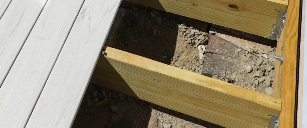 Close-up of wooden joists secured with metal brackets near the ledger board, with light gray composite decking being installed on top.