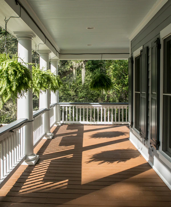 Covered Deck Builder in Council Bluffs, IA A traditional porch designed by a Covered Deck Builder in Council Bluffs, IA, featuring classic white columns, brown wooden decking, hanging fern plants, and dark window shutters.