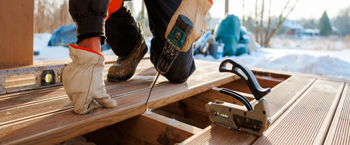 A worker in winter clothing kneels in the snow to drill into grooved boards during a cold weather deck repair, with a specialized deck spacing tool nearby.