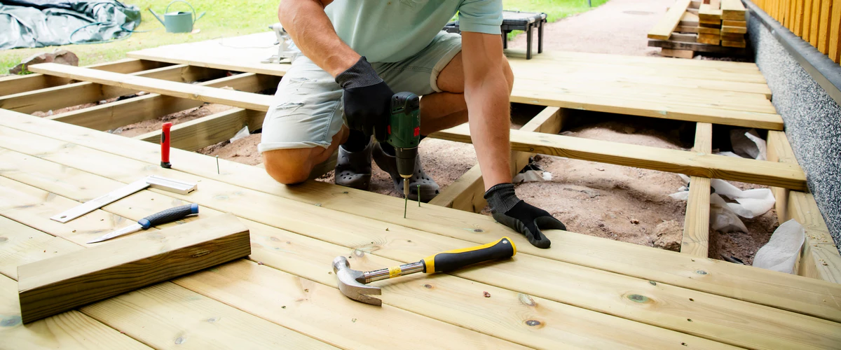 A worker performing a wood deck repair, kneeling in gloves to drill screws into new framing with a hammer, square, and utility knife resting nearby.