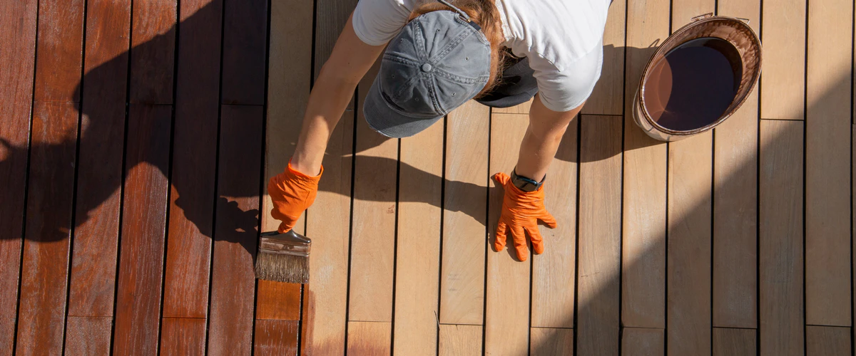 Top-down view of a person wearing orange gloves using a wide brush to apply a fresh coat of brown stain and sealant to a wooden deck.