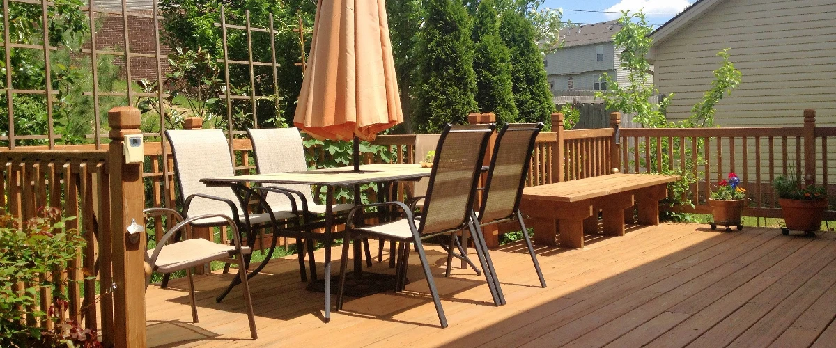A sunny backyard wooden deck furnished with a patio dining table, closed tan umbrella, matching chairs, and a built-in wooden bench next to garden greenery.