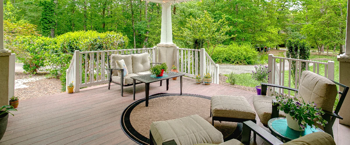 An inviting covered deck or porch with comfortable cushioned outdoor chairs, loveseat, ottomans, a central woven area rug, and numerous potted flowering plants, looking out onto a lush green wooded backyard.