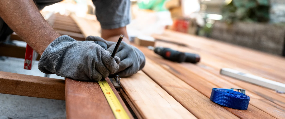 Close-up of a worker performing deck repair by meticulously measuring wood planks with a yellow tape measure and pencil for a perfect fit.
