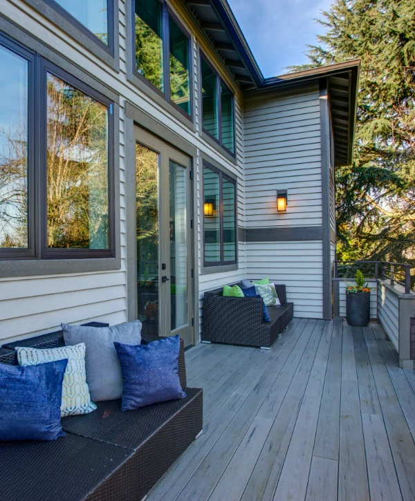 Grey composite deck installation in Fremont, NE showing a dark wicker outdoor sofa with blue and green throw pillows against a house exterior.