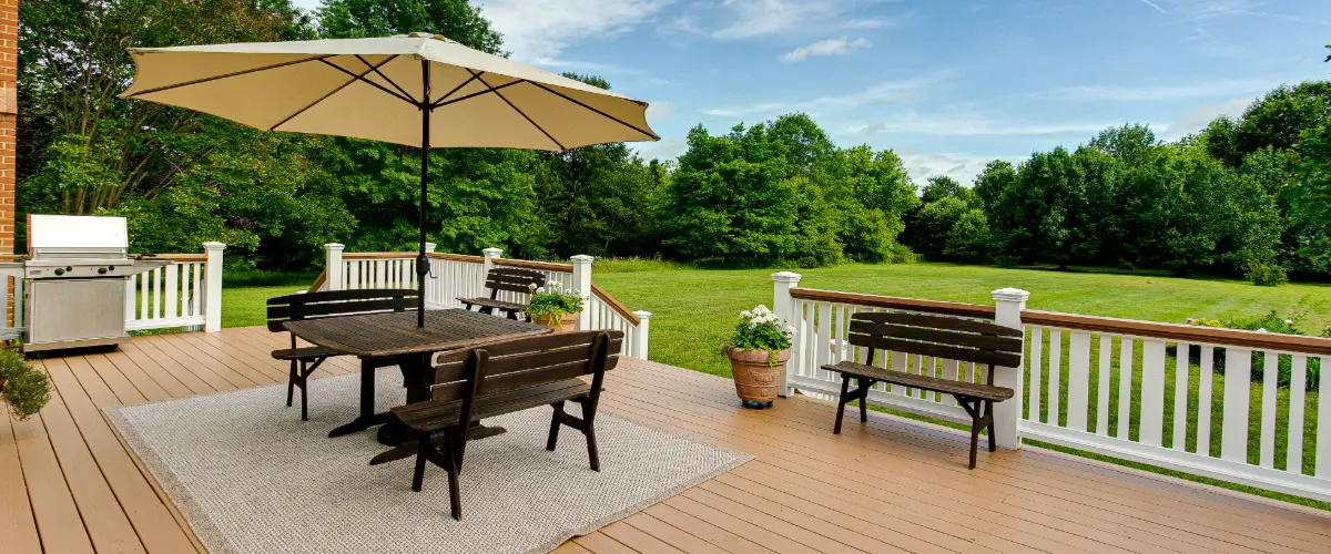 Spacious backyard wooden deck featuring a dining table with benches, a large beige umbrella, a grill, and white railings overlooking a green lawn.