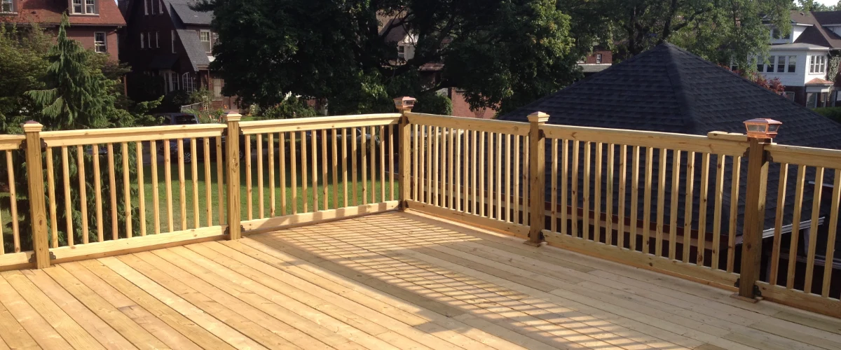 Natural wood deck featuring custom wooden railings with decorative copper post caps overlooking a residential neighborhood in bright sunlight.