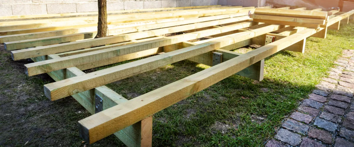 Treated lumber being used to build a solid deck frame, showing wooden joists and metal brackets laid out over a grassy yard during construction.