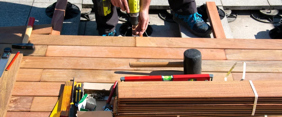 A close-up view of a contractor using a cordless power drill to screw down fasteners during a wooden deck renovation.
