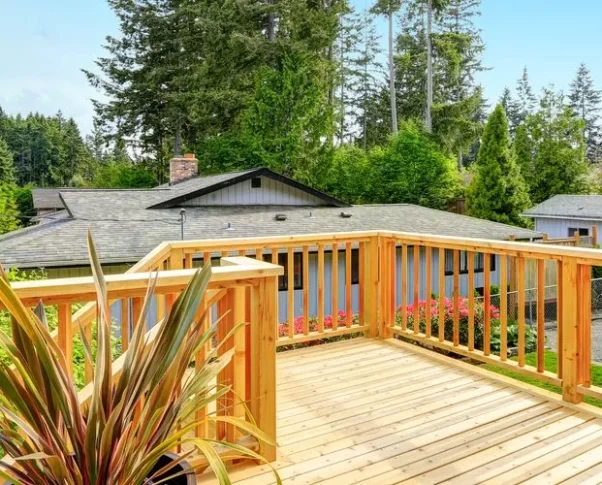 A wide-angle view of a professionally constructed elevated wooden deck made of light-colored cedar, featuring a sturdy vertical slat railing system overlooking a lush green backyard with tall evergreen trees and a neighboring house.